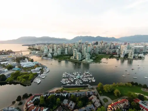 Aerial view of a coastal Vancouver city at dusk, showing the marina with boats, green parks, and the mountain range in the background.