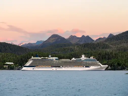 A Celebrity cruise ship anchored on the forested coastline against the backdrop of mountains and a pink-tinted sunset ready for an Alaskan cruise