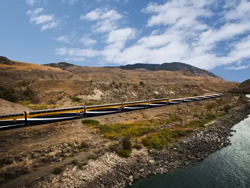 f Fraser Canyon with its dry, hilly terrain and the Rocky Mountain train under a bright blue sky.