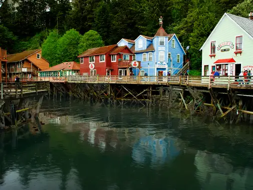 The quaint coastal village of Ketchikan , colorful wooden buildings on stilts along a pier, surrounded by lush green trees in a small coastal village.
