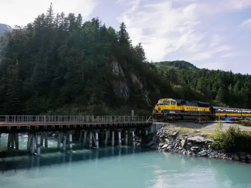 A scenic view of Holland America Line Cruisetrain crossing a wooden bridge over a turquoise river, with lush green trees in the background.